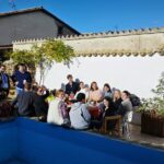 Grupo de personas en un porche de una casa de pueblo, varias personas sentadas al rededor de una mesa y varias levantadas.