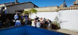 Grupo de personas en un porche de una casa de pueblo, varias personas sentadas al rededor de una mesa y varias levantadas.